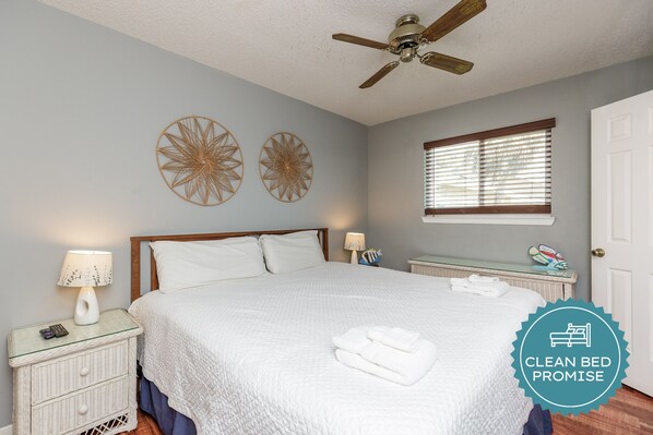A clean, well-lit bedroom with a bed, two wall decorations, a ceiling fan, a window with blinds, and a bedside table with a lamp.