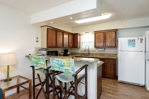 A kitchen with wooden cabinets, a white refrigerator, a microwave, and a coffee maker. There is a small table with a lamp and two bar stools with patterned cushions.