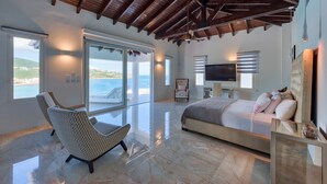 A spacious bedroom with a large bed, a sitting area with a patterned chair, a television, and a view of the sea through large windows. - Villa Amalia (Philipsburg)