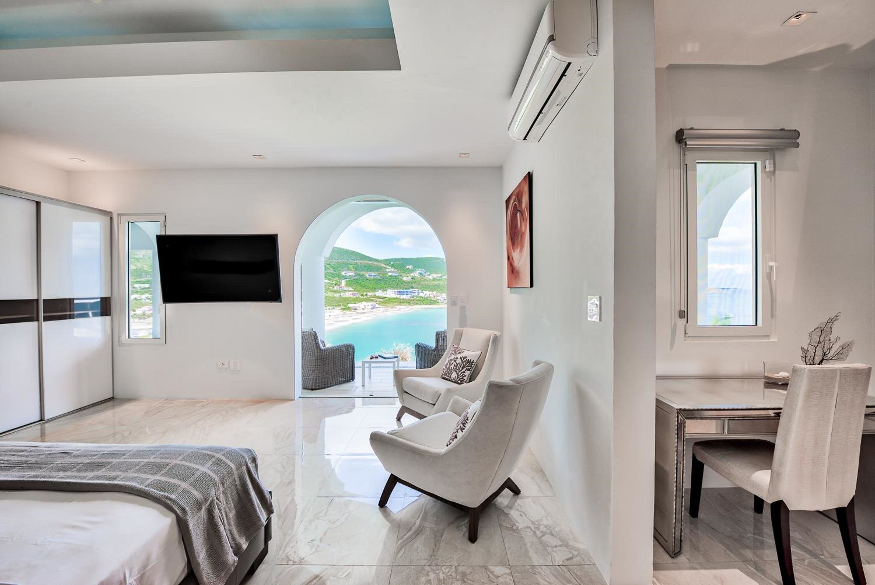 A modern bedroom with a bed, a desk, and a chair. There is a large window with a view of a beach and a mountain.