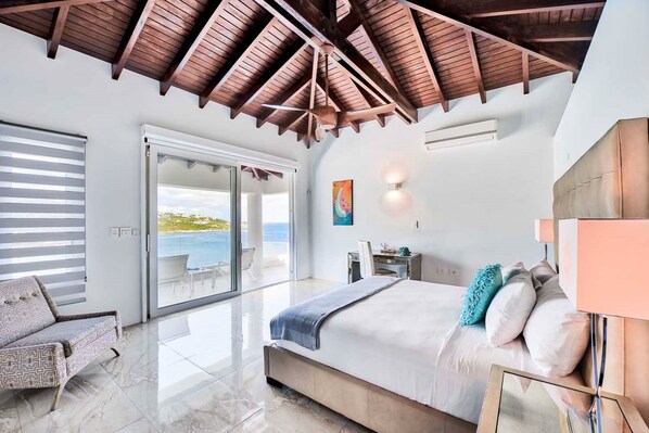 A bedroom with a large bed, a sofa, a desk, and a view of the sea through a sliding door. - Villa Amalia (Philipsburg)