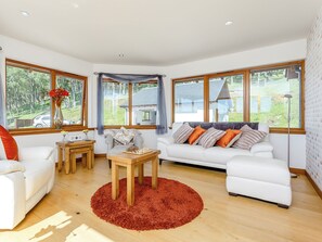 A modern living room with a white sofa, a wooden coffee table, and large windows offering a view of the outdoors. - 3 bedroom accommodation in Carrbridge, near Aviemore (Carrbridge, near Aviemore)
