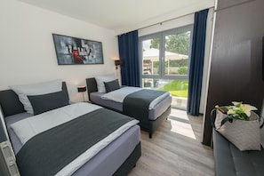 Una habitación de hotel con dos camas, un ventanal con cortinas, un cuadro en la pared y una mesita de noche con una lámpara.