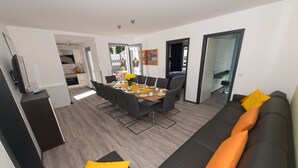 A modern living room with a dining area, black and orange sofas, a wooden floor, and a kitchen visible through an open door.
