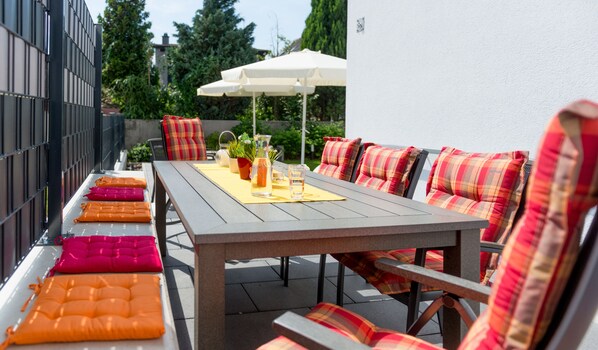 Eine Terrasse im Freien mit einem Holztisch, mehreren Stühlen und einem weißen Sonnenschirm.