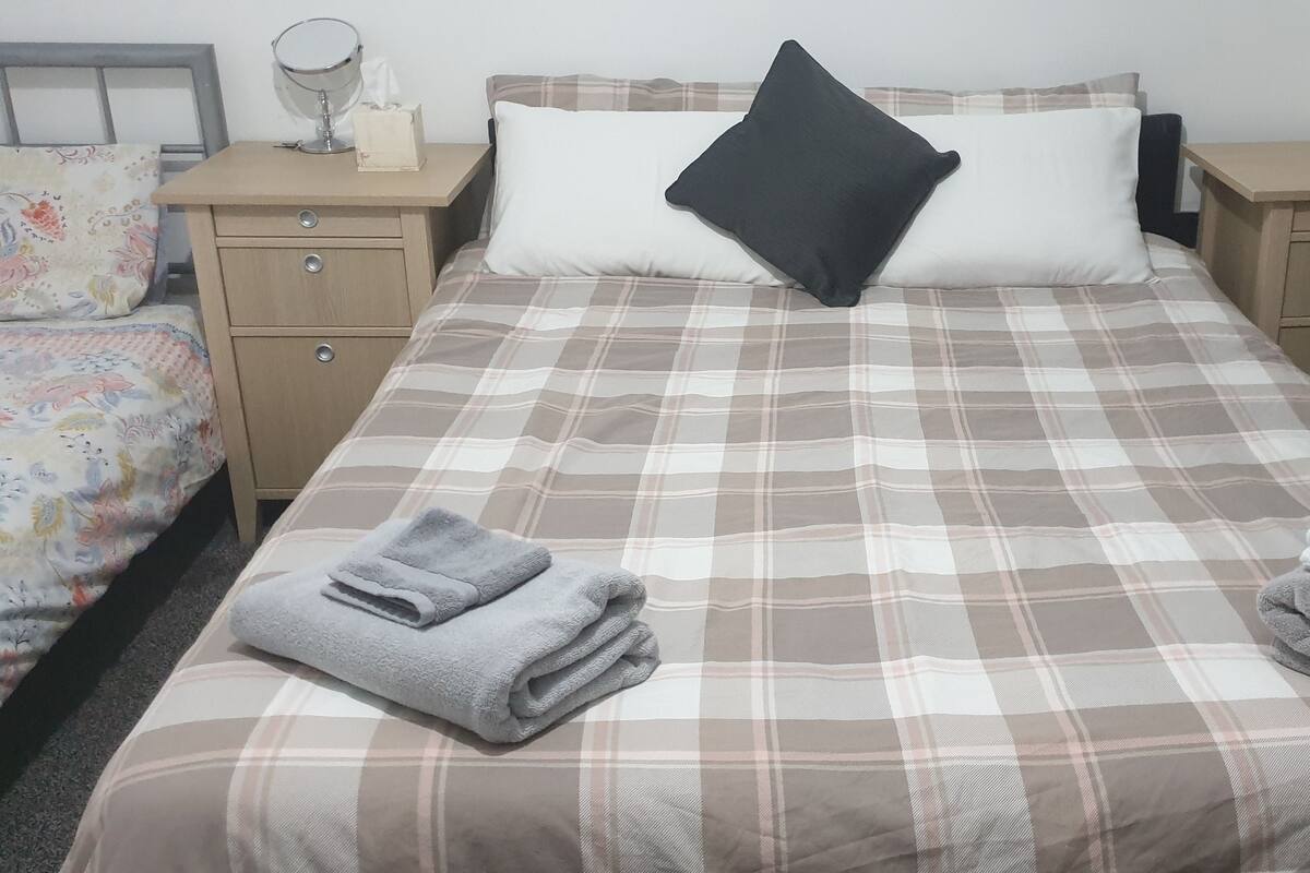 A neatly made bed with a plaid bedspread, two bedside tables with lamps, a mirror, and a small nightstand with a tissue box.