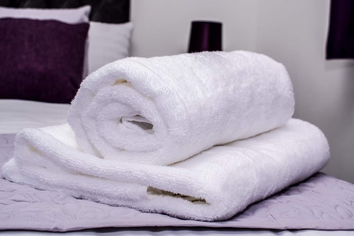 A neatly folded white towel stacked on a bed with a purple bedspread.