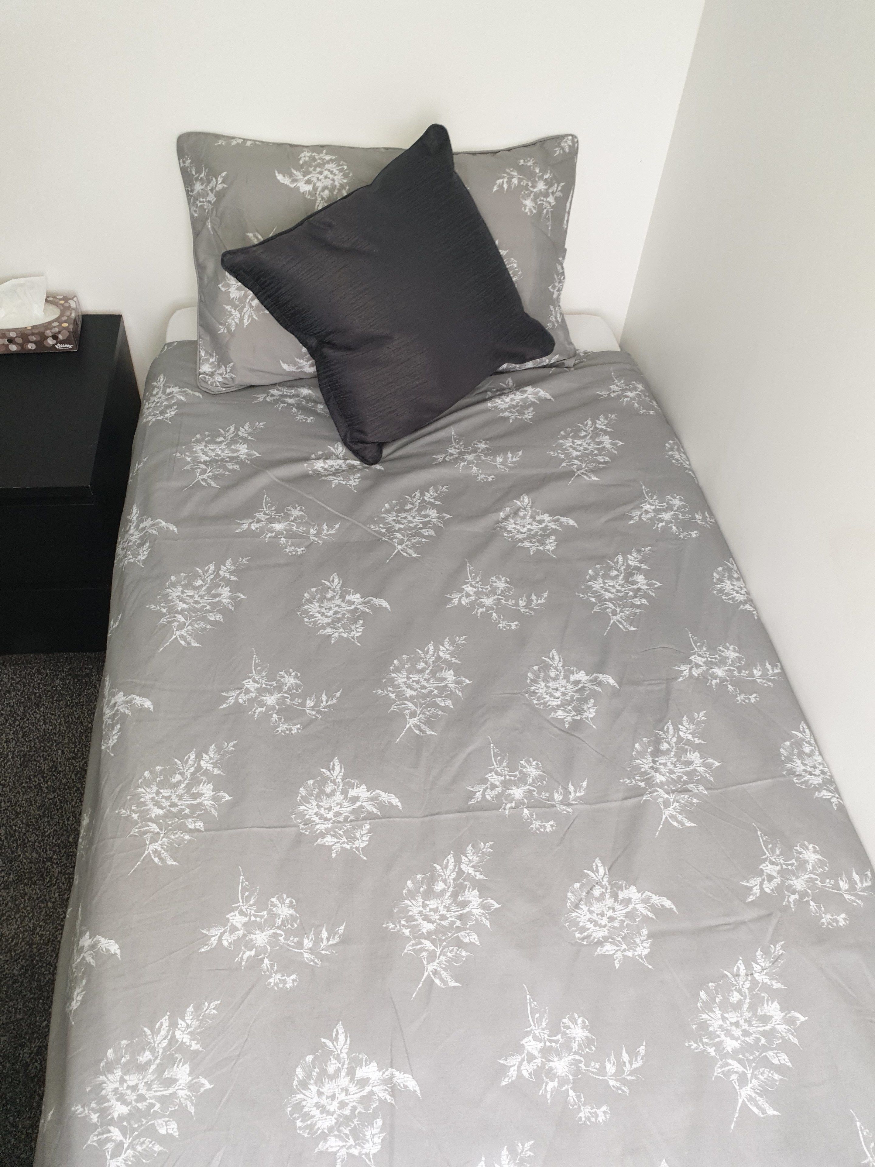 Una cama individual con una colcha gris estampada con flores y una almohada negra.