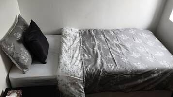 A small, neatly made bed with floral bedding and a dark pillow, positioned against a plain wall with a window on the right.
