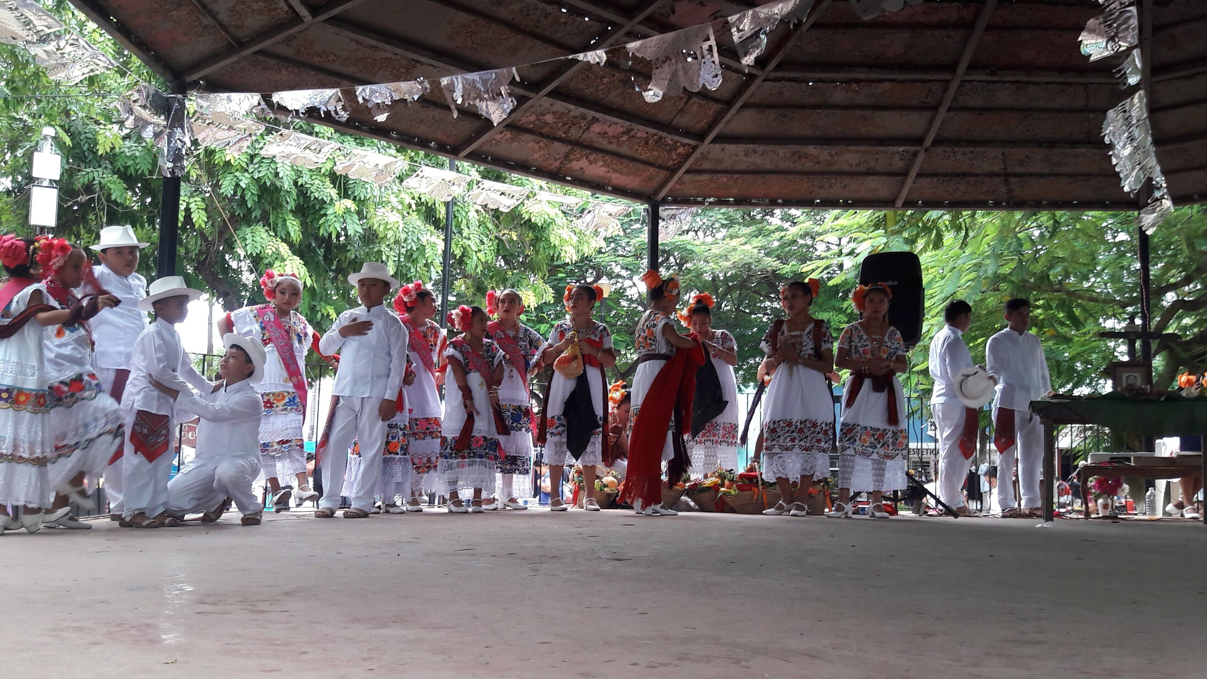 Un groupe de personnes, vêtues de costumes traditionnels, se trouve sous une zone abritée dont le sol est en béton.