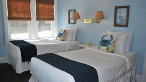 Quarto de hotel com duas camas, uma janela com persianas, luminárias fixadas na parede e um quadro emoldurado.