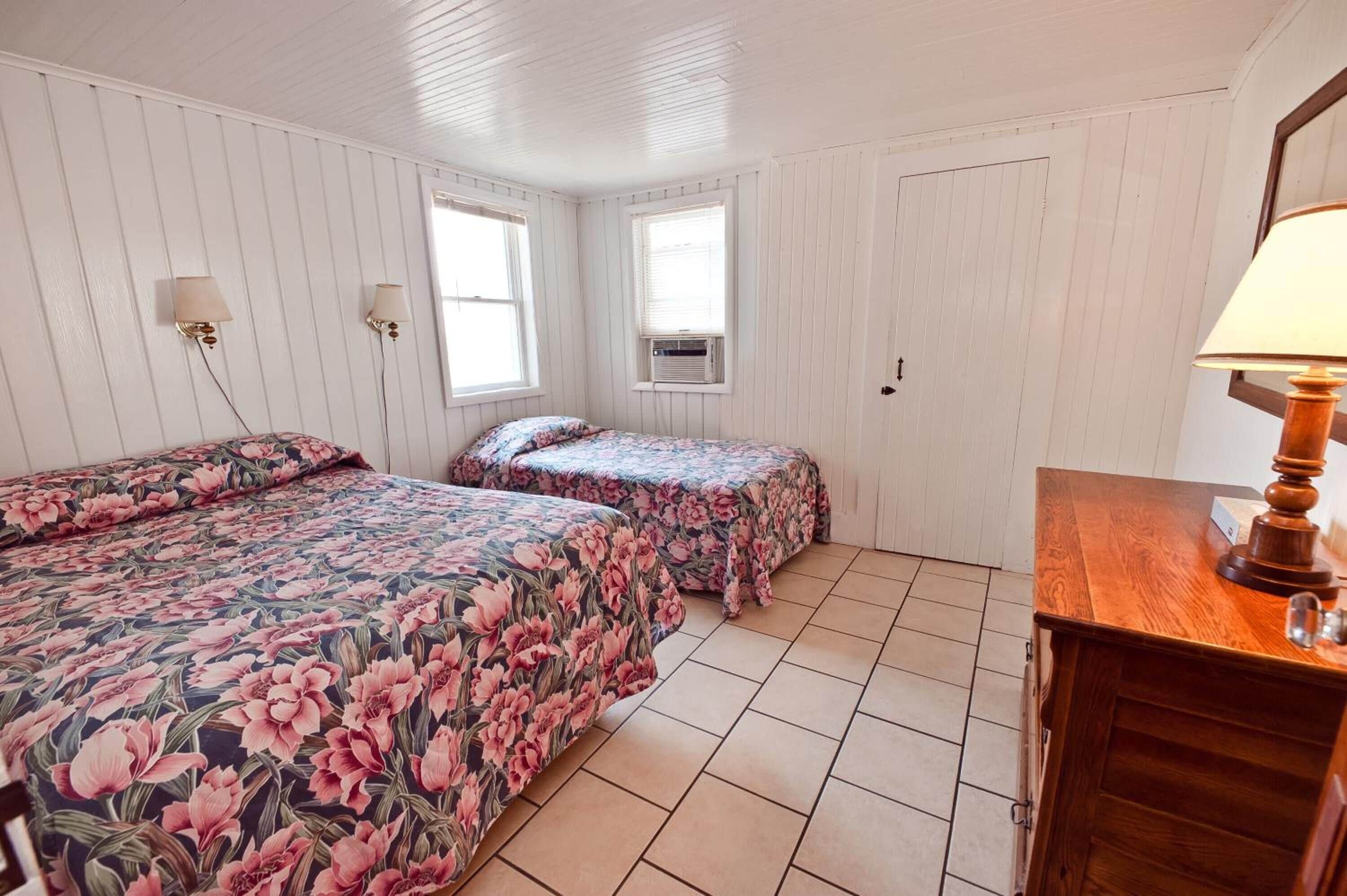 Una habitación con dos camas, paredes blancas, un aparador de madera y dos lámparas de pared.