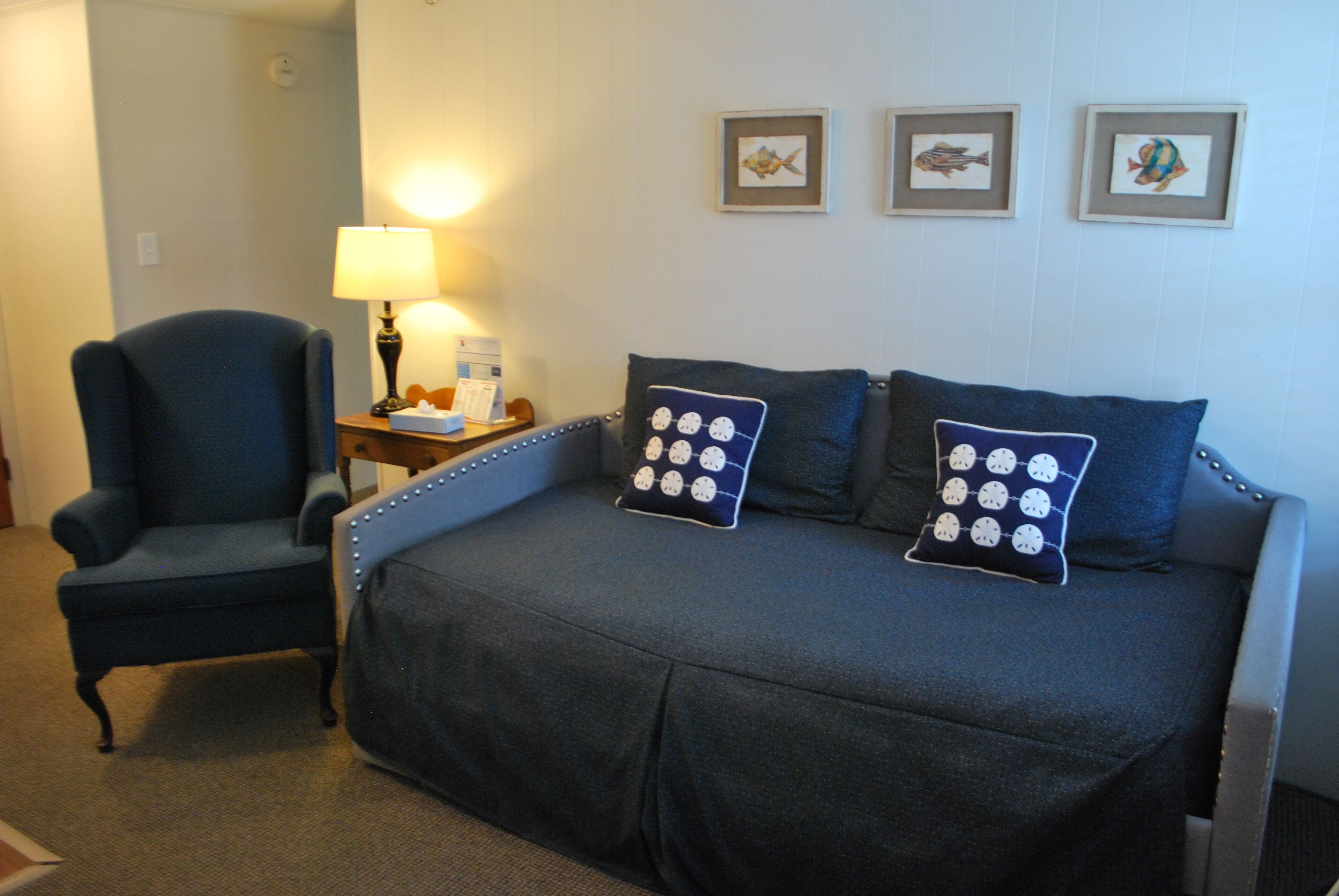 A hotel room with a dark blue sofa bed, a matching armchair, a small table with a lamp, and three framed fish pictures on the wall.