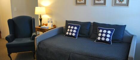 A hotel room with a dark blue sofa bed, a matching armchair, a small table with a lamp, and three framed fish pictures on the wall.