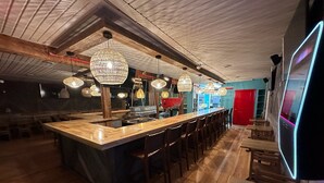 Uma área de bar com um balcão de madeira comprido, várias luminárias pendentes e uma porta vermelha ao fundo.