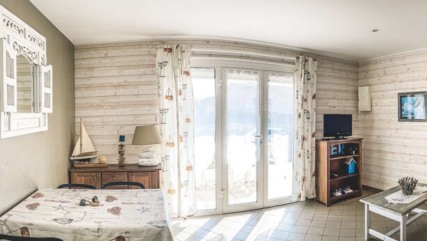 Una sala de estar acogedora con un muro de madera, una mesa de comedor con mantel estampado, un sofá, una mesita de centro y un televisor sobre un mueble de madera.