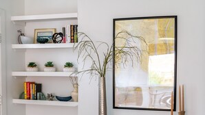 A room with white walls, a wooden ceiling, a glass console table with books, a framed abstract painting, and a potted plant. - Palmetto Cottage - Historic 1925 cottage in Downtown Fairhope (Fairhope)