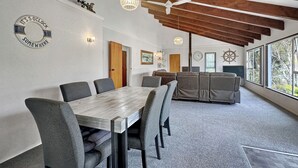 A modern dining area with a wooden table and grey upholstered chairs, a ceiling fan, and a large window with a view of trees. - Central Spacious Family House - Paihia House (Paihia)