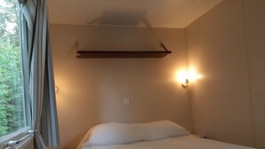 Uma cama de solteiro com roupa de cama branca num quarto com janela, mesa de cabeceira e prateleira na parede.