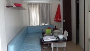 Quarto pequeno e moderno com sofá azul, mesa de jantar com bandeja, um guarda-chuva vermelho e janela com cortinas.
