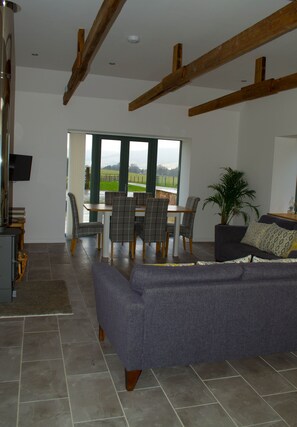 A spacious living room with a large grey sofa, a dining area with a table and chairs, and a view of the outdoors through large windows.