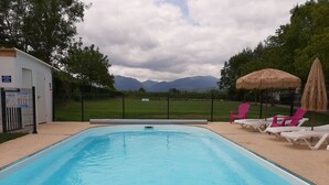 A swimming pool with lounge chairs and a thatched umbrella, surrounded by a fence and trees, with mountains in the background. - Camping Uhaitza - Chalets (Gotein-Libarrenx)