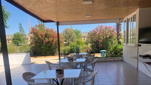 Un patio al aire libre con techo de madera, sillas de plástico blanco y una mesa. Hay un televisor montado en la pared y se ve una vista de árboles y edificios al fondo.