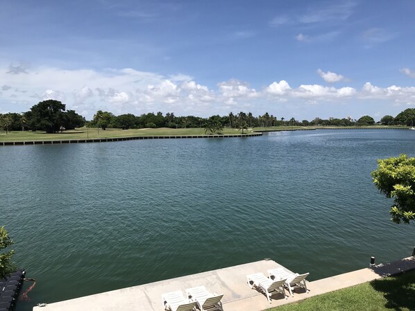 A view of a calm lake with a grassy area and trees in the background. There are lounge chairs on a concrete platform in the foreground.