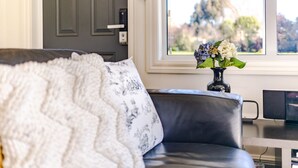 Un salon avec un canapé gris, un plaid blanc à motifs, un vase avec des fleurs, une fenêtre donnant sur des arbres et une porte avec un interrupteur.
