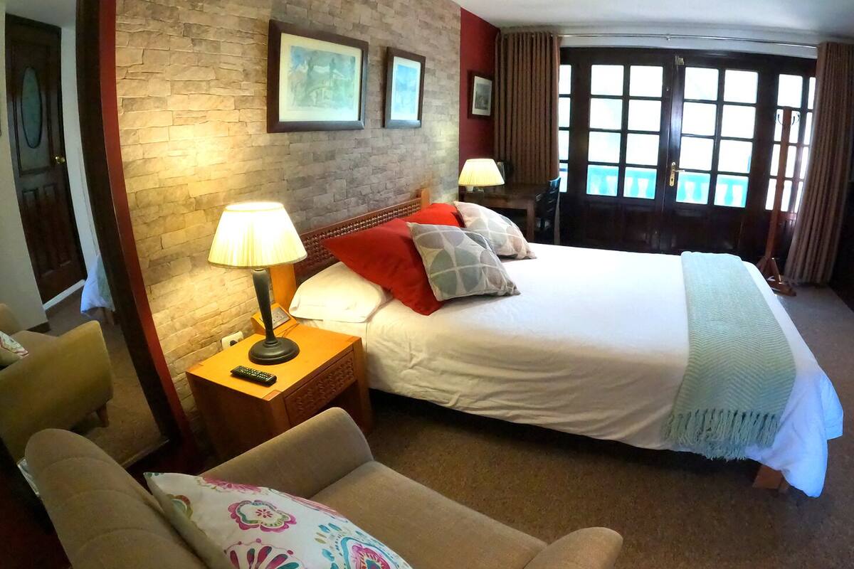 A hotel room with a large bed, a nightstand with a lamp, a chair, and a desk with a computer.