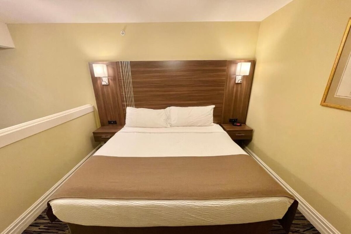 A hotel room with a large bed, wooden headboard, two bedside tables with lamps, and a carpeted floor.