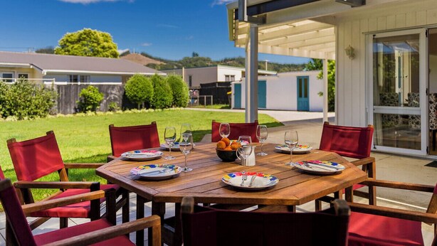 Zona pranzo all'aperto con tavolo di legno e sedie rosse, con sullo sfondo un quartiere residenziale e un cielo sereno.
