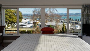 A bedroom with large windows offering a view of a cityscape and water. - Jacaranda Lodge - Napier Hill Holiday Home (Napier City)