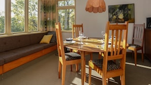 A dining area with a wooden table, chairs, and a sofa. There are windows with curtains, an air conditioning unit, and a painting on the wall. - Jacaranda Lodge - Napier Hill Holiday Home (Napier City)