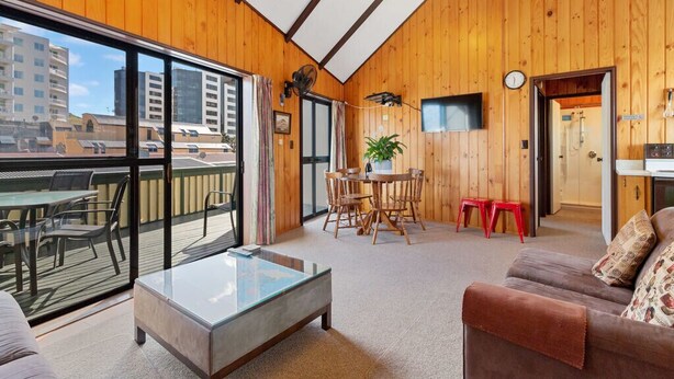 A spacious room with wooden walls, a glass coffee table, a sofa, a dining area with a table and chairs, and a view of buildings through large windows.
