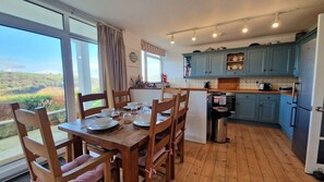 A dining area with a table set for four, a kitchen with blue cabinets, and a view of the countryside through large windows. - 2 Steepfield, spacious apartment, Bantham S. Devon (Bantham)