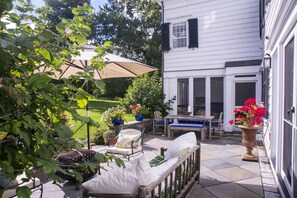 Une terrasse avec des meubles d’extérieur, une table et des chaises, entourée de plantes en pot et d’un grand parasol.