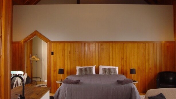 A bedroom with wooden paneling, a bed with gray bedding, two bedside tables with lamps, and a chandelier. - Revamped church in an old gold mining village (Central Otago District)