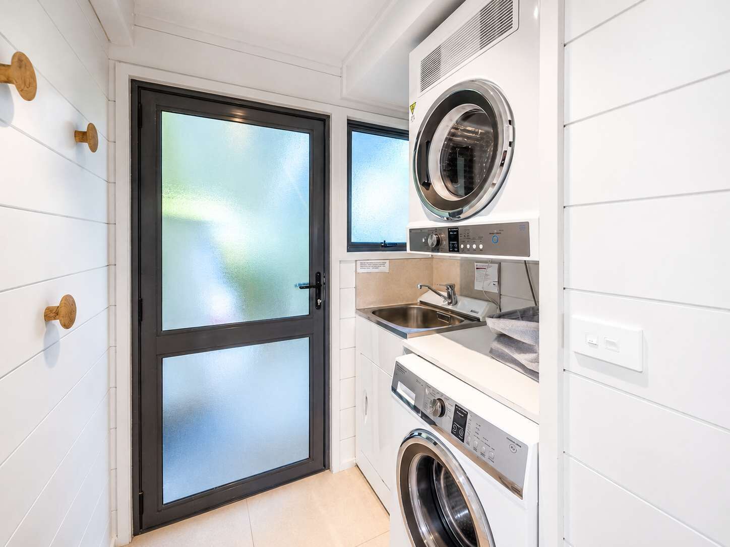 A compact laundry room with a front-loading washer and dryer, a sink, and a frosted glass door.