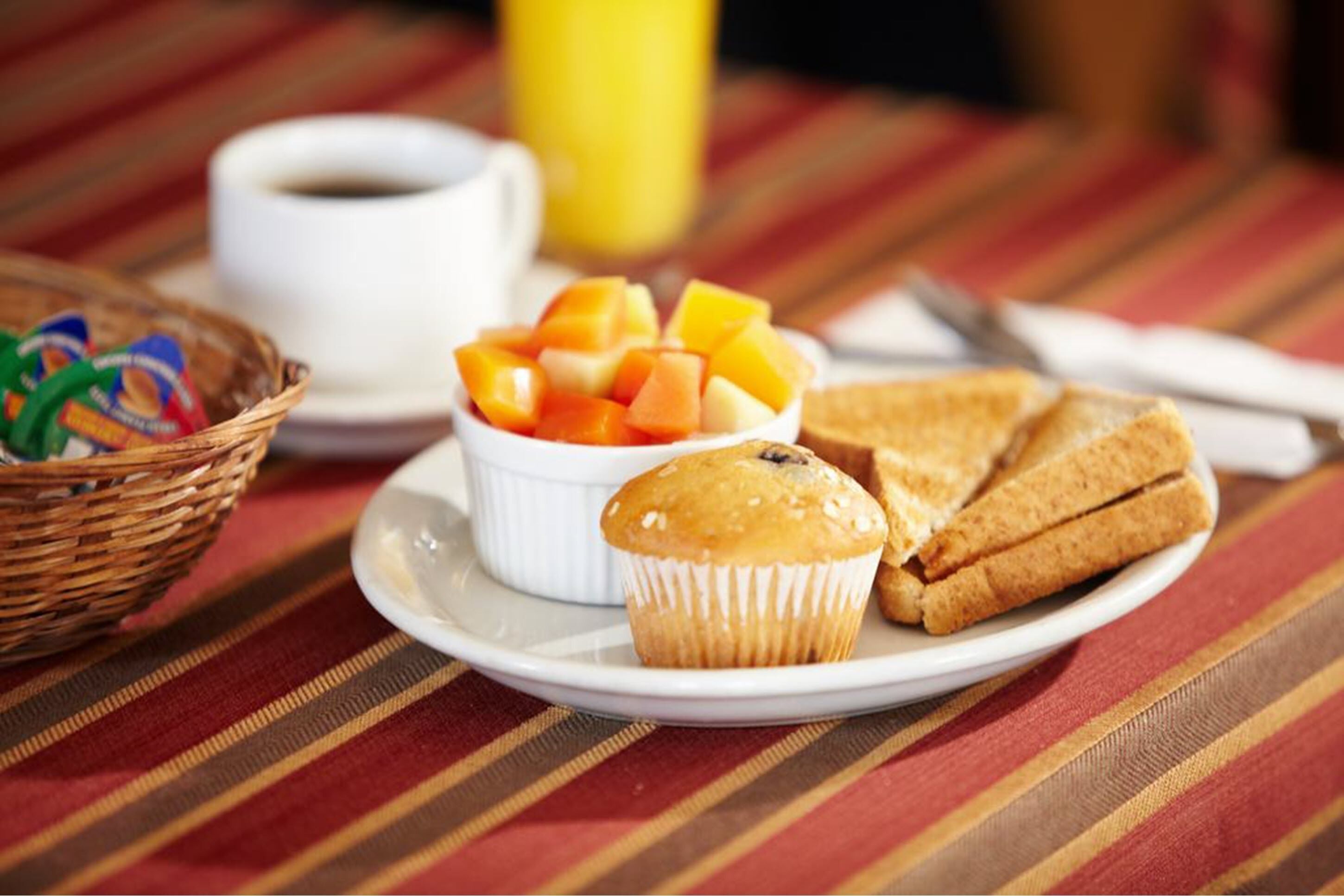 A breakfast setting with a plate of muffins, toast, and fruit, a cup of coffee, and a basket of bread.
