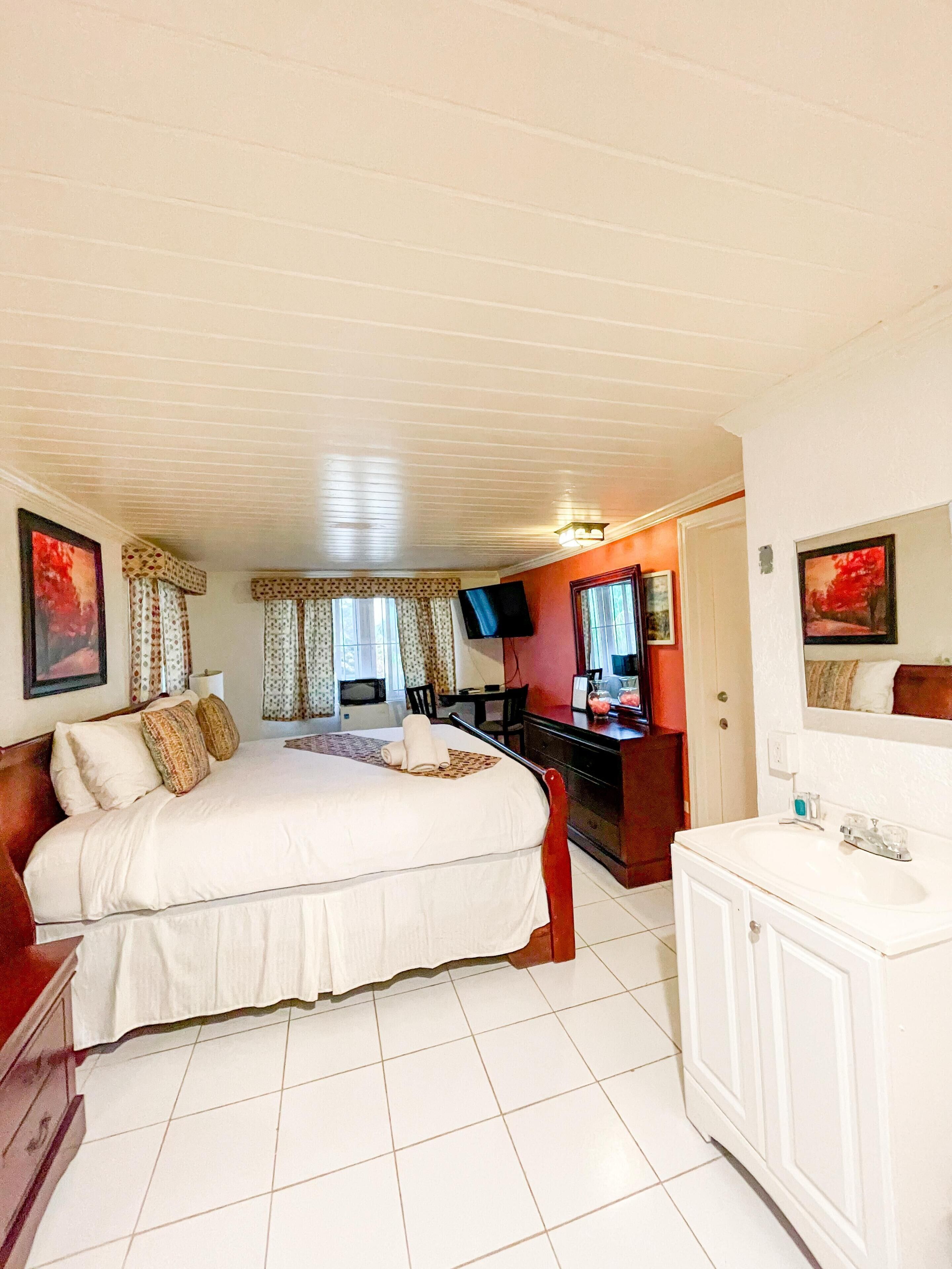 A hotel room with a large bed, a desk, a TV, and a bathroom with a sink and mirror.