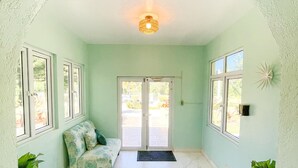 Un pasillo amplio con pared verde claro, techo blanco y un gran arco de entrada a un espacio exterior. Se observa un banco con cojín estampado, una puerta con panel de vidrio y un adorno en la pared.