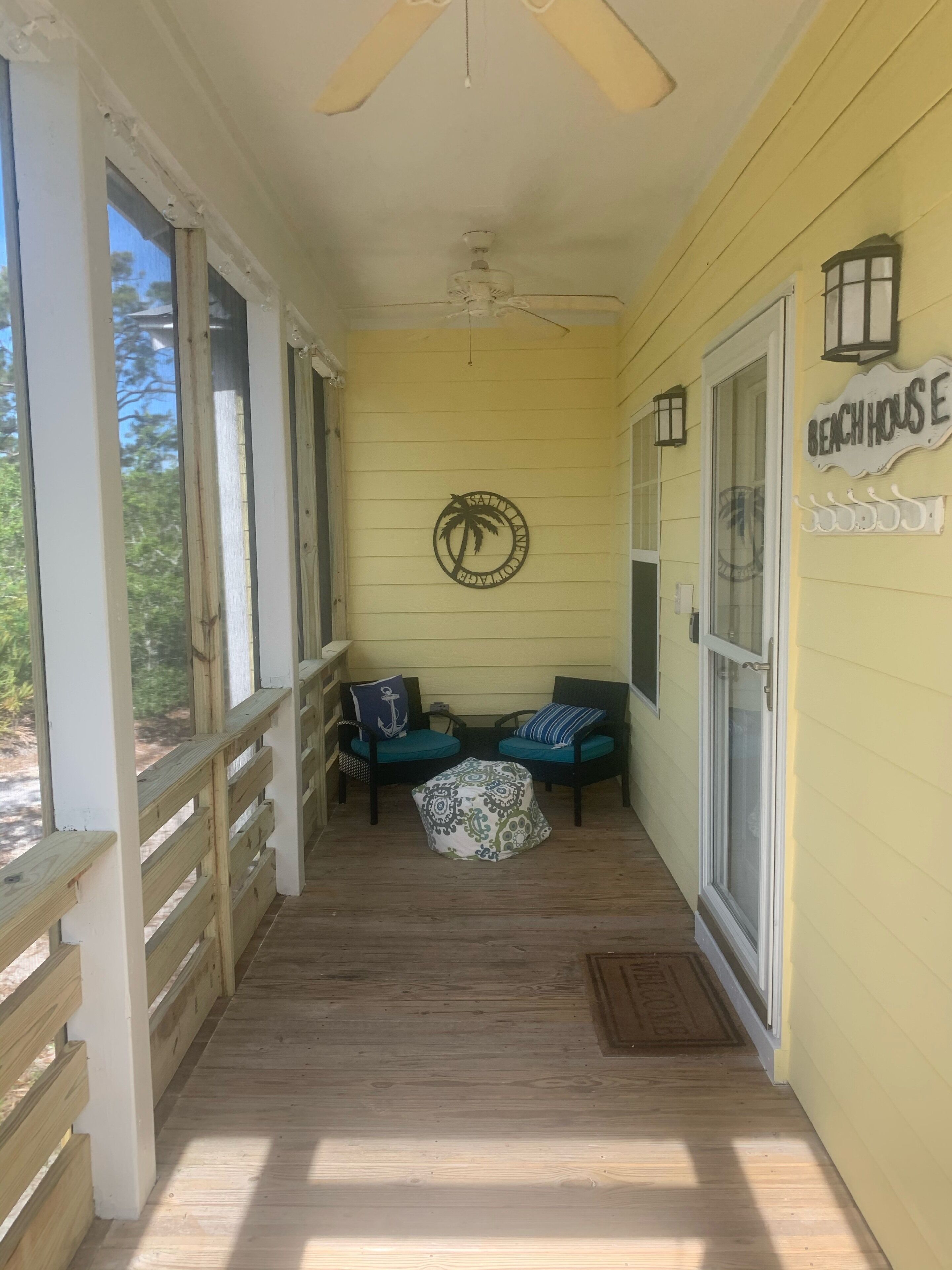 Une véranda couverte avec deux chaises et une petite table, un ventilateur de plafond et une pancarte indiquant « Maison de plage ».