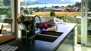 A modern kitchen with a large window offering a view of a coastal town and mountains. - Enjoy the view (Waikawa Bay)