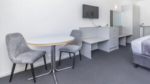 A modern hotel room with a round table, two grey chairs, a white desk with drawers, a flat-screen TV mounted on the wall, and a bed with grey bedding.
