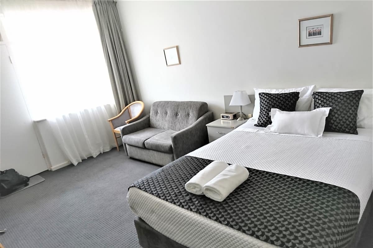 Quarto de hotel com cama, poltrona, mesa de cabeceira, abajur e um quadro na parede.