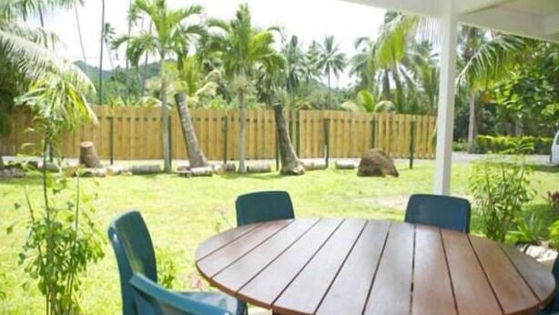 Een houten tafel met vier stoelen in een overdekt buitengebied, met een houten hek en palmbomen op de achtergrond.