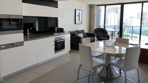 Une cuisine moderne avec des meubles blancs, un four encastré et un four à micro-ondes. On y trouve un coin repas avec une table et des chaises blanches, ainsi qu’une grande fenêtre offrant une vue sur la ville.