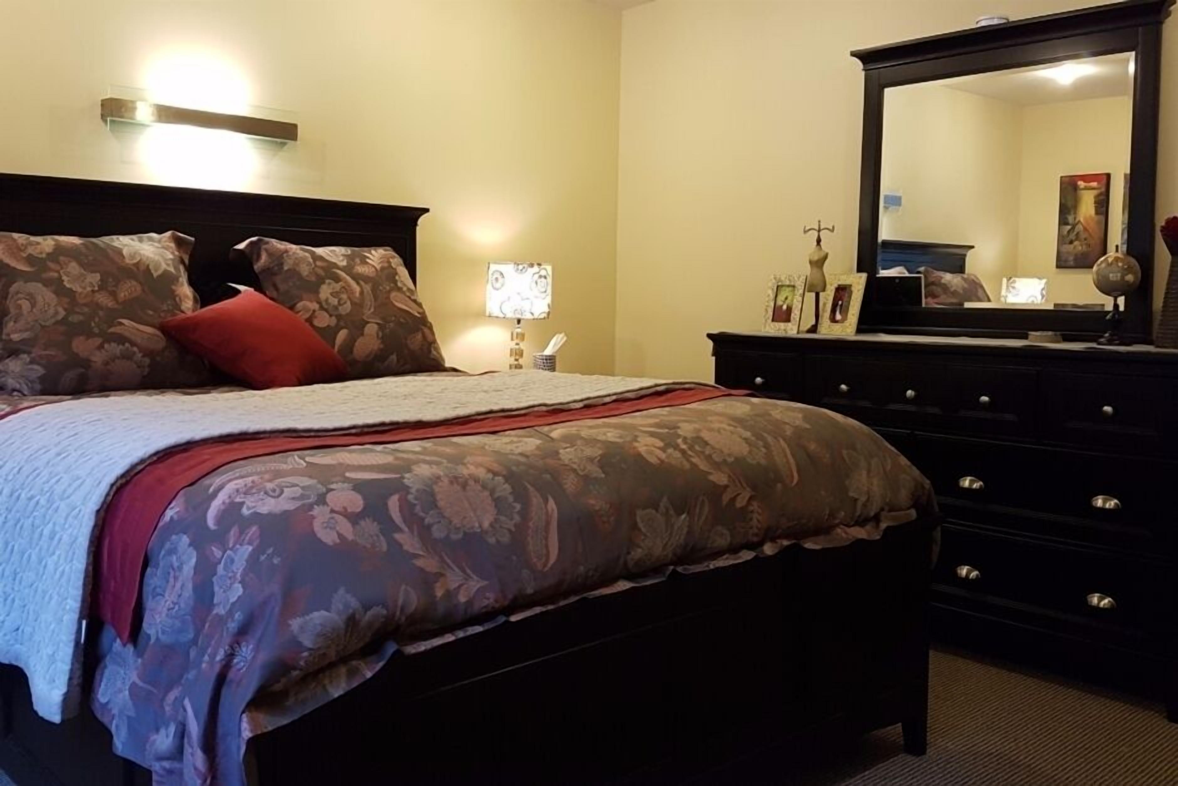 A neatly made bed with floral bedding and a red pillow, a dresser with drawers, a mirror, and a wall-mounted lamp.