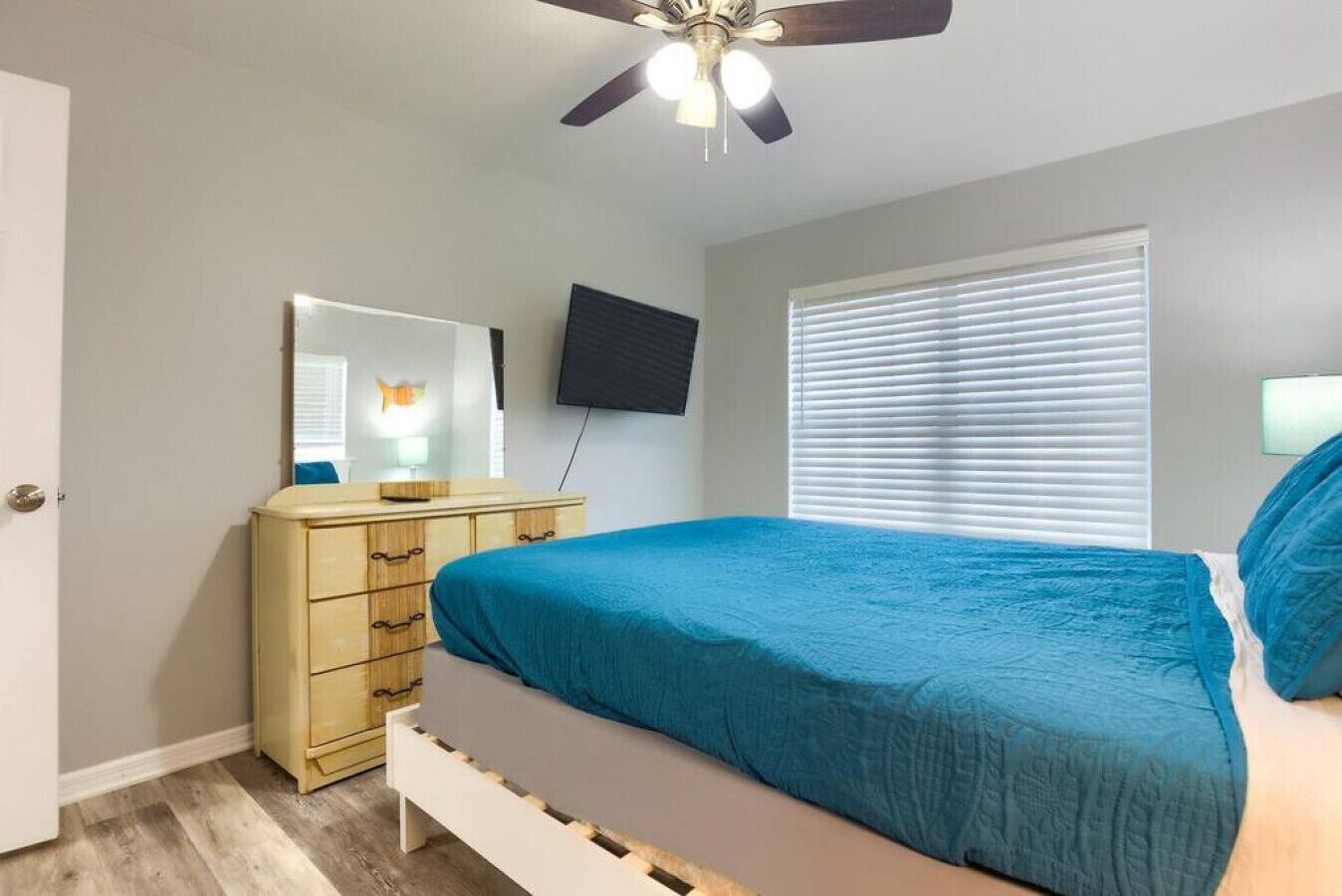 Un dormitorio con cama, cómoda, ventilador de techo, televisor y ventana con persianas.